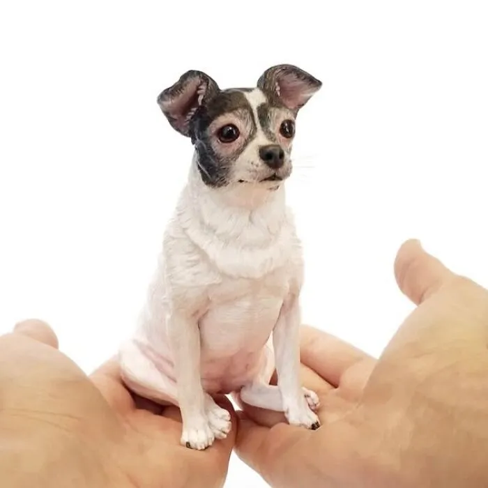 Person holding a tiny, handmade hyperrealistic sculpture of a small chihuahua-like dog.