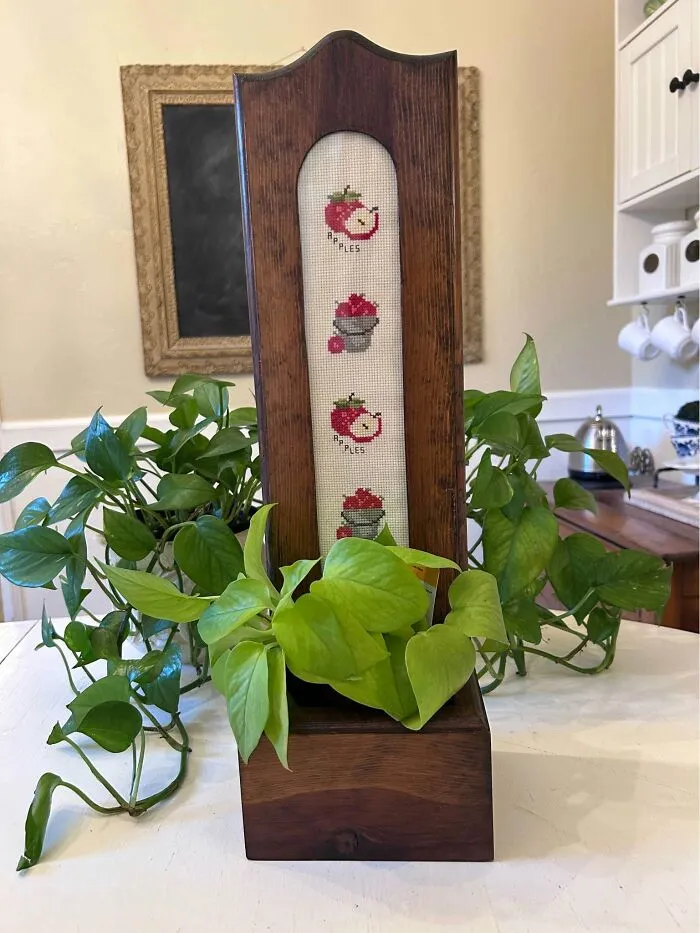 Wooden planter with cross-stitched apple design and green plant inside thrift store find
