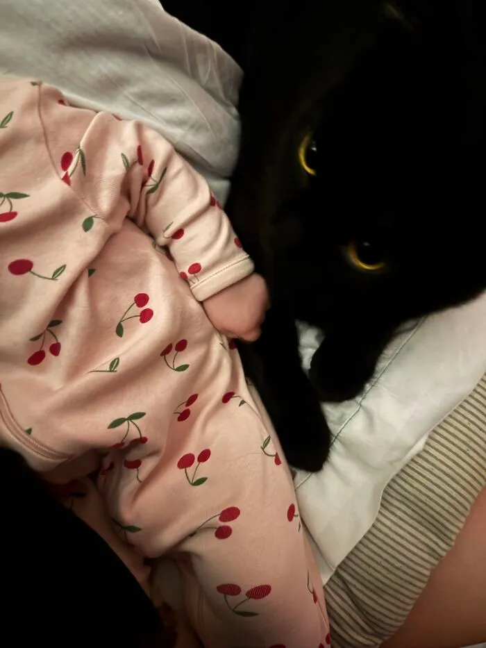 A black cat resting closely next to a baby wearing cherry-patterned pajamas