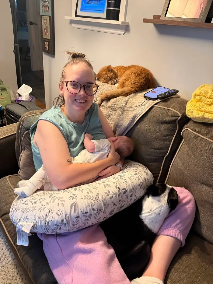 A mother holding her newborn baby on the couch surrounded by family pets looking after babies