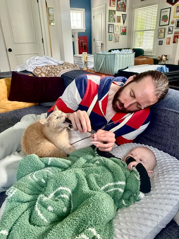 A man filing a cat's nails while relaxing on the couch next to a newborn baby