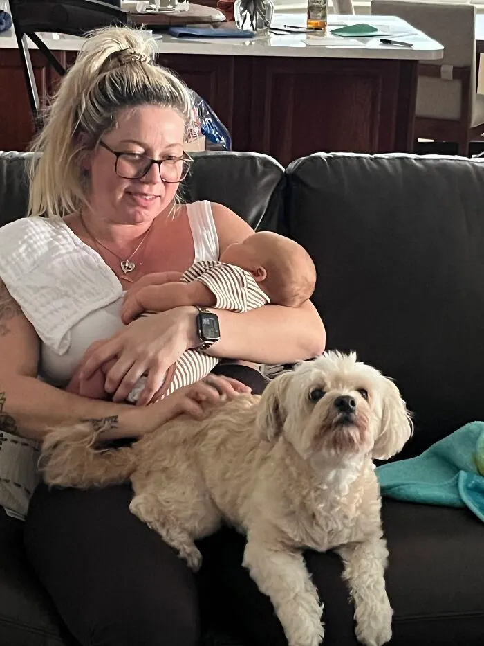 Mother holding a newborn while petting a small white dog sitting on the couch