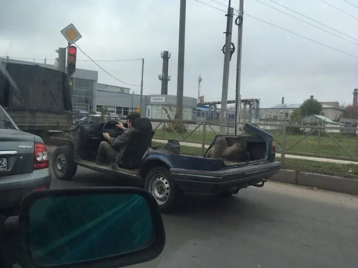 A person driving a stripped-down car shell in traffic, demonstrating bizarre Slavic engineering.