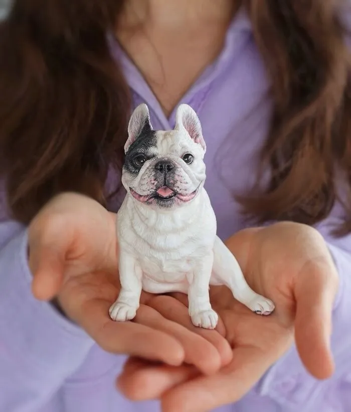 A hand holding a hyperrealistic sculpture of a French bulldog pet.