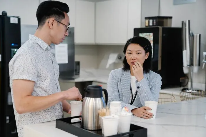 Two coworkers casually talking over coffee in office kitchen admitting disturbing things