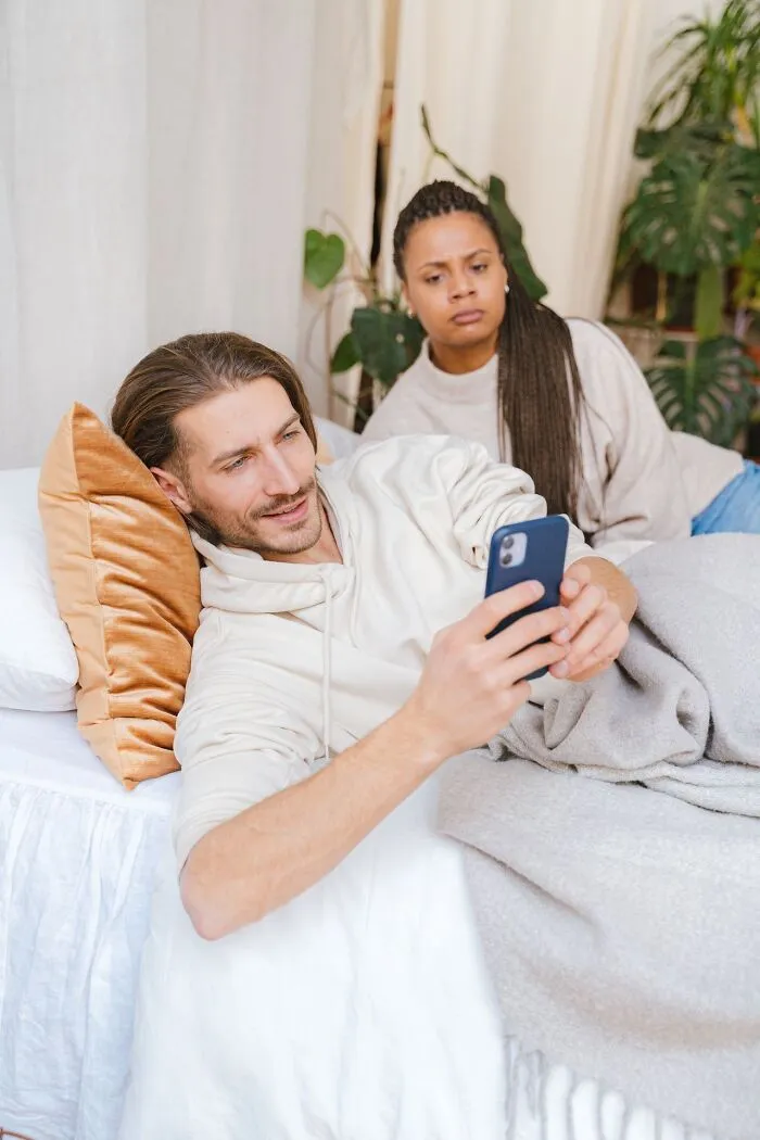A woman looking suspicious at a man using his phone in bed, highlighting reasons to choose divorce.