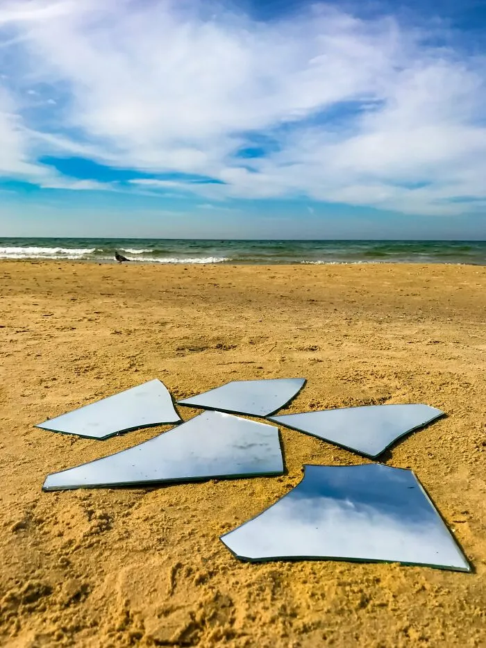 Broken mirror pieces reflecting sky on beach sand with ocean background