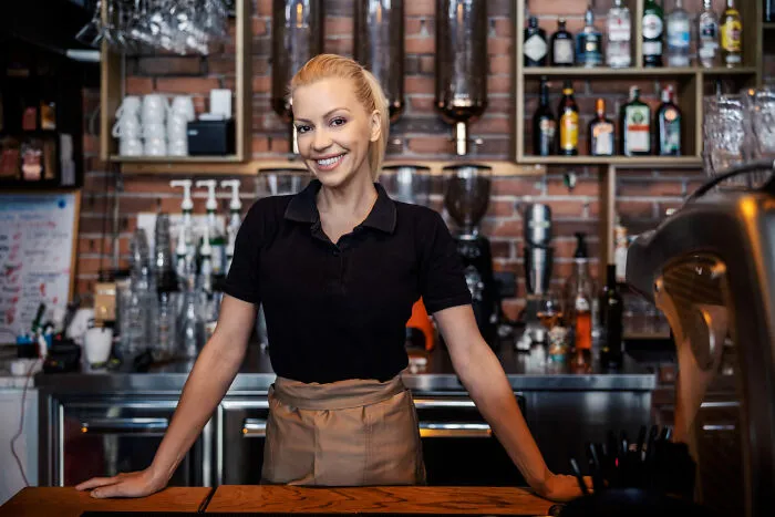 A friendly female barista smiling behind a coffee shop counter while listning to what strangers said.