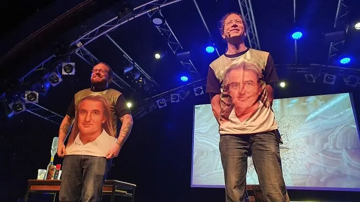 Two men on a stage wearing printed face shirts, performing in front of an audience about unusual cults