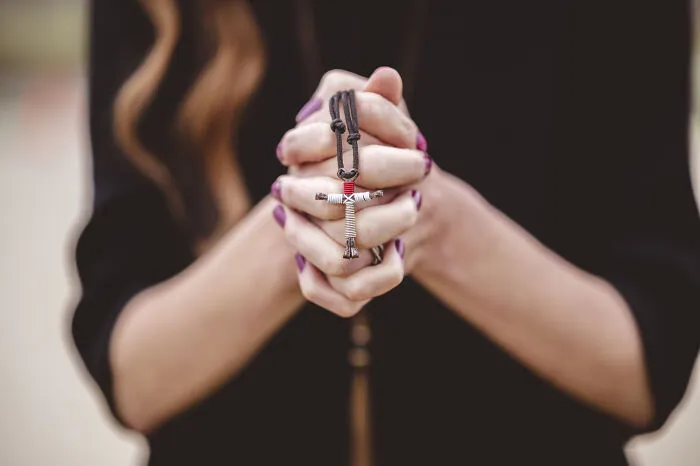 Close-up of hands clasped in prayer holding a cross necklace, referencing lesser-known cults.