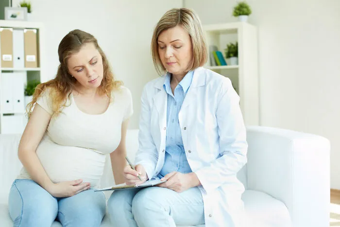 A female doctor writing on a clipboard while consulting with a pregnant patient.