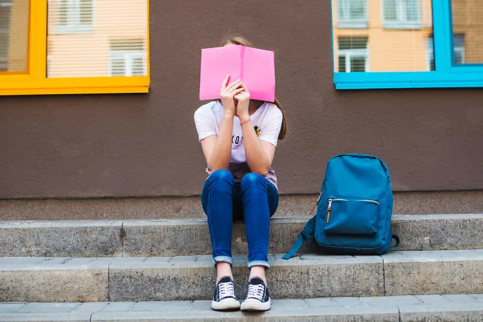 A young female student sitting outside holding a pink notebook, trying to hide from infuriating teachers