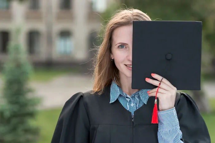 A proud female graduate holding her graduation cap over her face, representing the pressure of helicopter parenting.