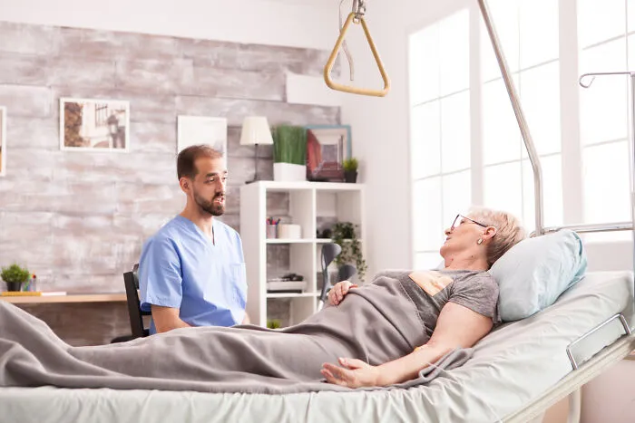 A male nurse in blue scrubs talking to an elderly female patient lying on a hospital bed.