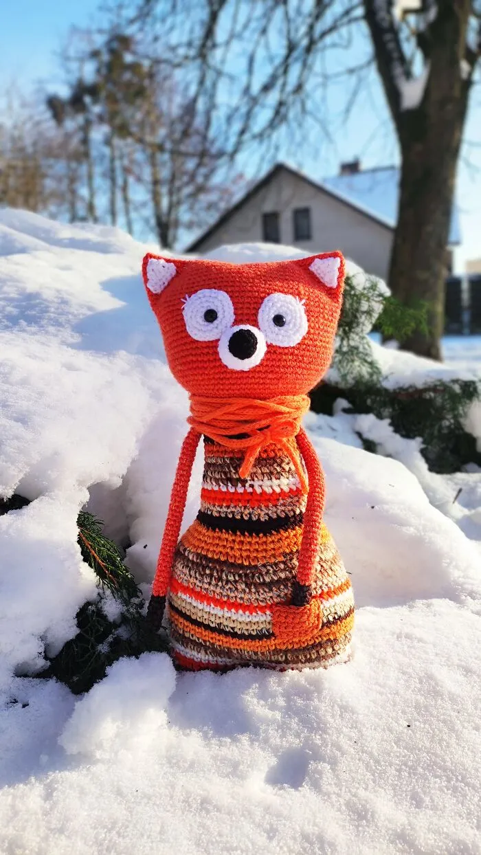 A red fox crochet creature wearing an orange scarf standing in the snow.