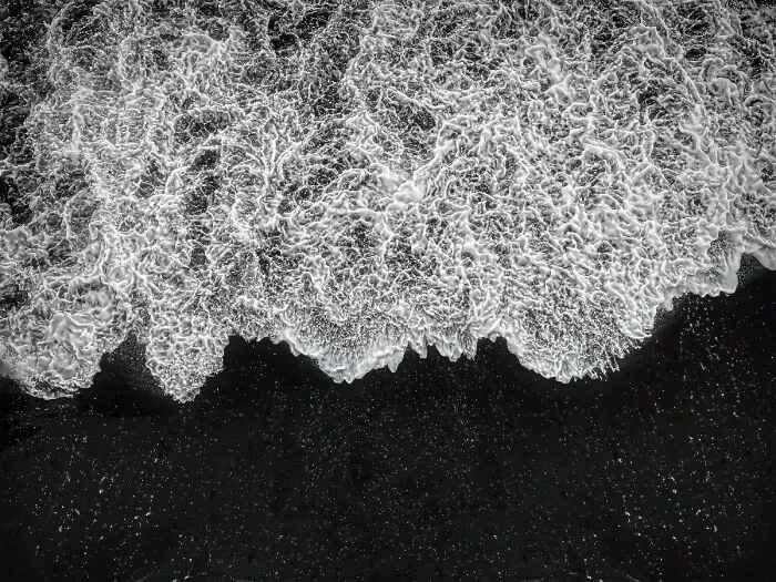 Aerial view black-and-white photo of ocean foam crashing against a dark shoreline.