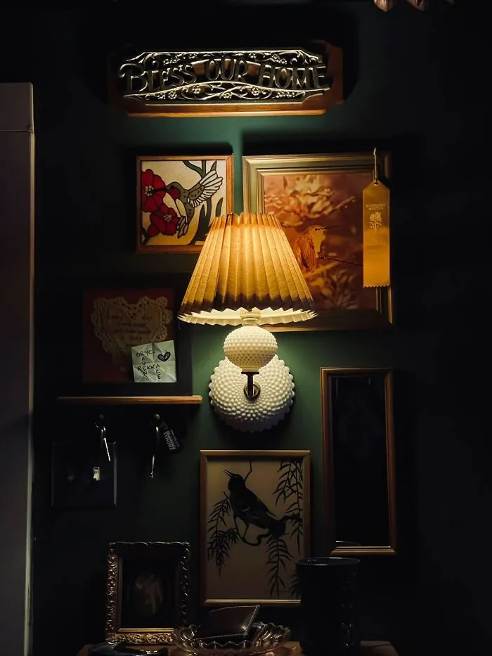 A lighted vintage wall lamp mounted on a dark green wall among framed art thrift finds.