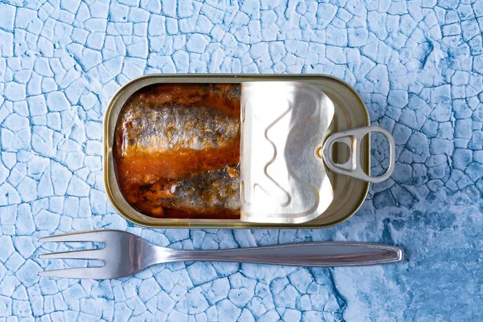 Open can of sardines with fork on textured blue surface for hilarious revenge
