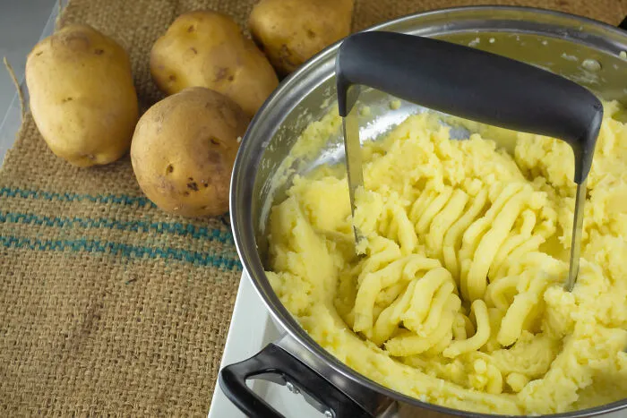 Potato masher in bowl of mashed potatoes with raw potatoes nearby for cooking humor