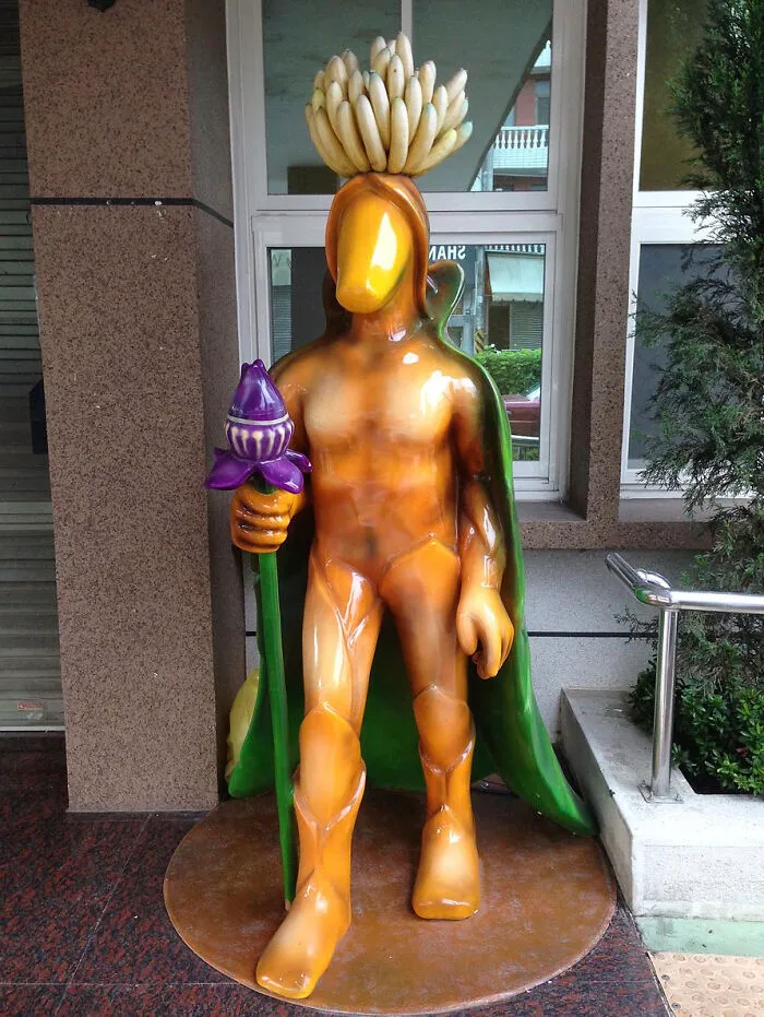 A bizarre orange human-shaped statue of public art featuring bananas on its head and holding a purple bud
