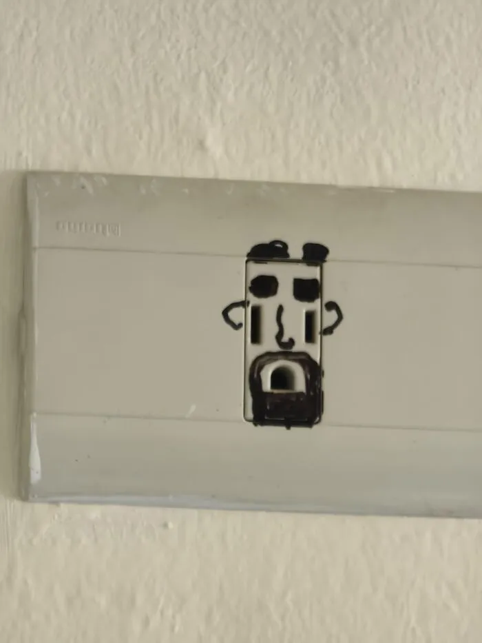 A wall power outlet drawn with marker to look like a face, creating an absurd moment.