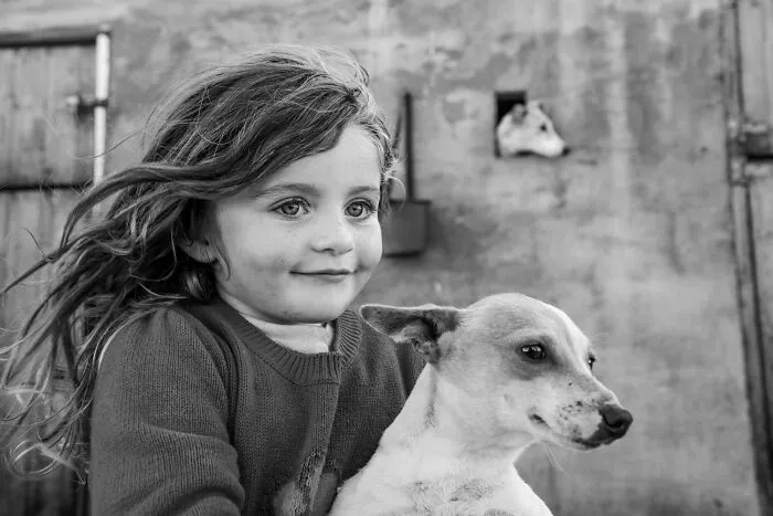 A black-and-white photo of a young girl smiling while holding her small dog outdoors.