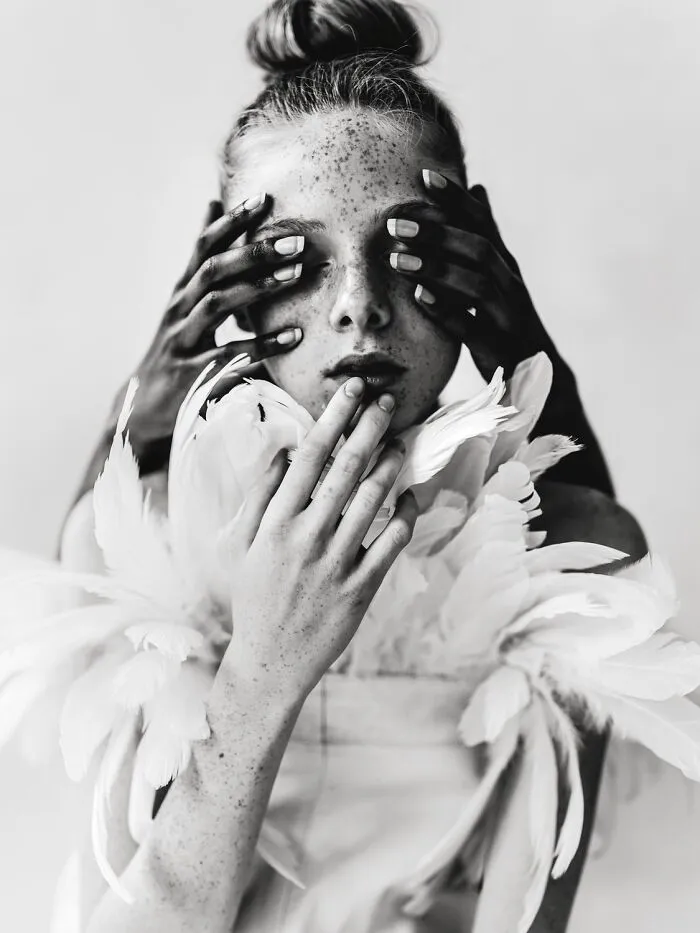 Artistic black-and-white photo featuring a woman with freckles, feather accents, and hands covering her eyes.