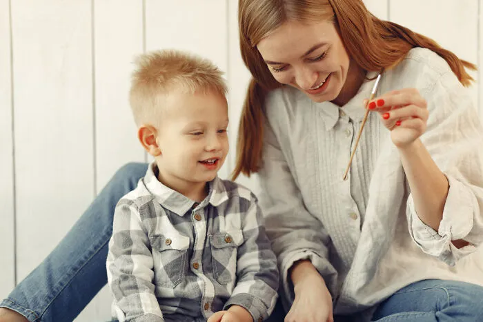 A smiling mother guiding her young son on how to paint, showcasing hands-on helicopter parenting style.