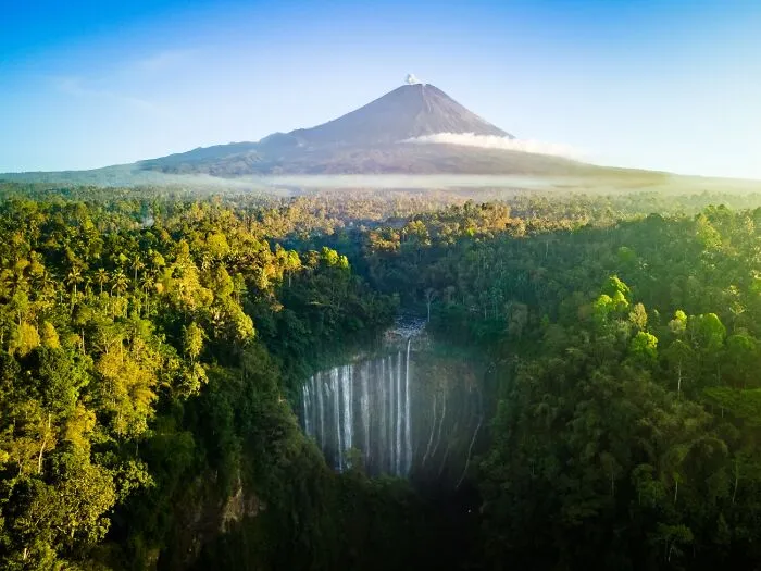 Lush tropical forest waterfall with a smoking volcano in the background from 2026 Siena Drone Photo Awards.