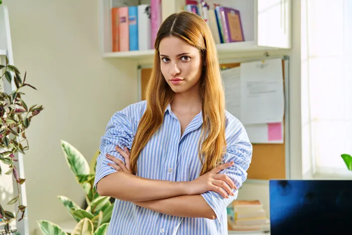 An infuriating teacher standing with crossed arms in a classroom setting, looking strict.