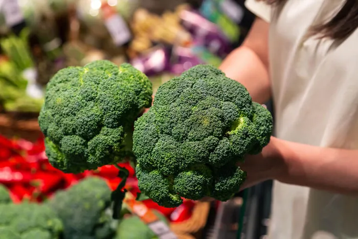 A person holding two heads of green broccoli while questioning partner's basic food knowledge