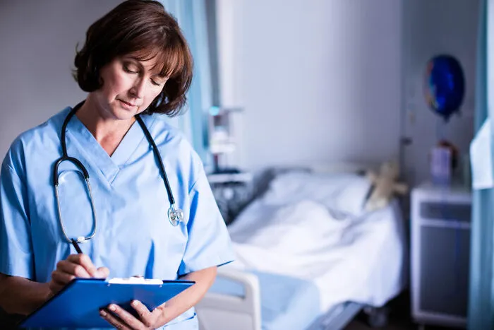 Nurse in blue scrubs holding a clipboard inside a hospital room.