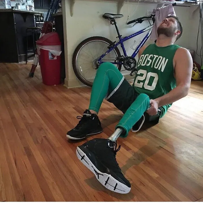 Man with prosthetic leg sitting on floor wearing Boston Celtics jersey as hilarious costume.