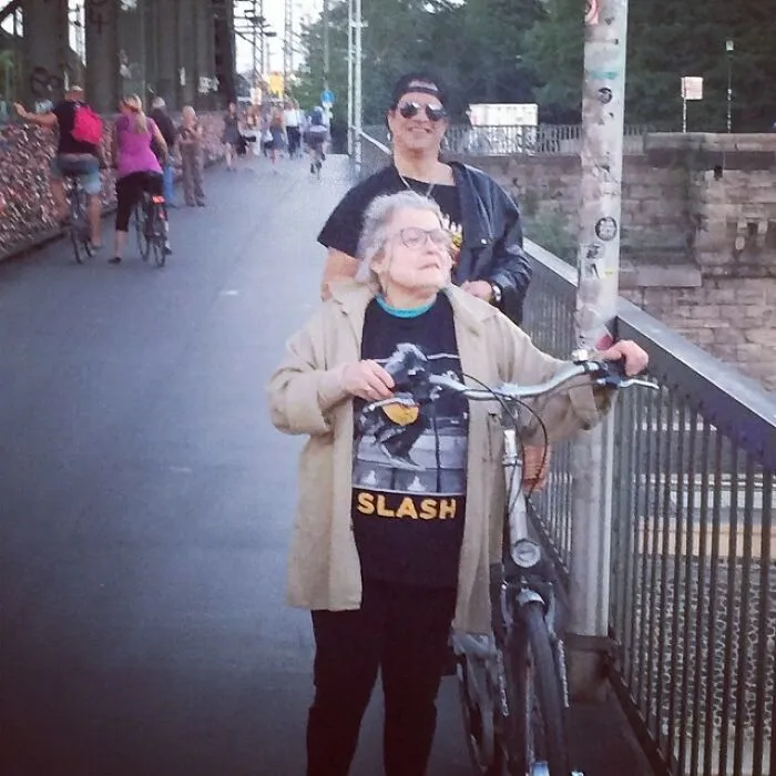 A man resembling Slash stands in the background behind an elderly woman wearing a Slash t-shirt