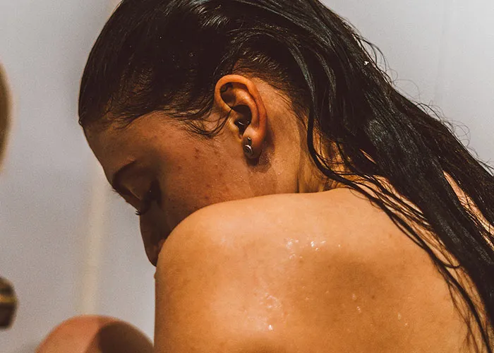A woman with wet hair looking down in the shower, depicting postpartum recovery after giving birth.
