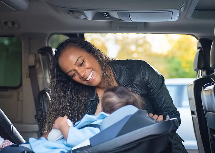 A smiling mother checking on her baby in a car seat, experiencing new motherhood.