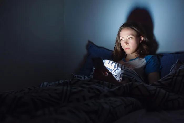 A young woman in bed in the dark looking at her phone, perfect for reading chilling scariest true stories.
