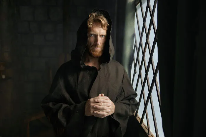Red-bearded man in a dark hood standing near a window, illustrating quick-fire jokes.