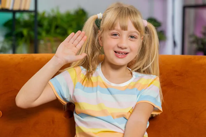 A smiling young girl with pigtails raising her hand sitting on an orange couch for quick-fire jokes