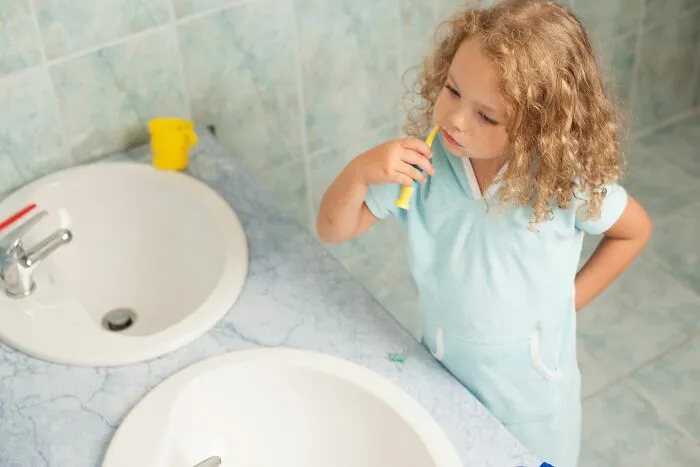A young blonde girl in a blue outfit brushing her teeth in a bathroom, reflecting unethical life pro tips