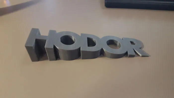 A grey 3D printer plastic door stopper spelling the word Hodor sitting on a desk