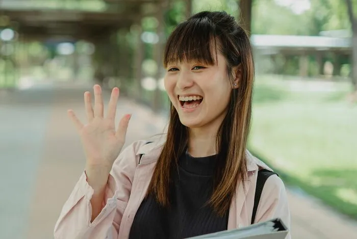 Smiling woman waving outdoors with greenery in background