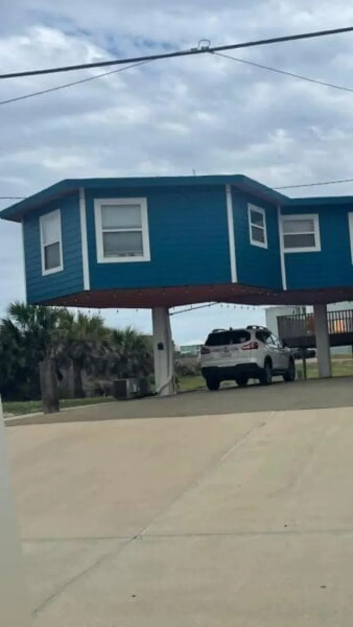 A blue house strangely elevated on thin stilts over a parking area, featured in cursed images.