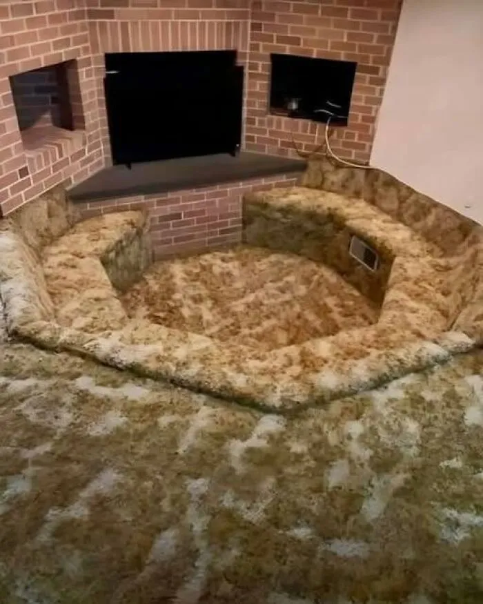 An interesting sunken conversation pit completely covered in brown carpet next to a brick fireplace.