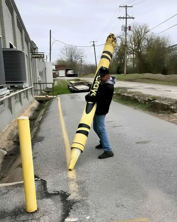 An odd photo of a man holding a giant yellow Crayola crayon on a road near a yellow line.