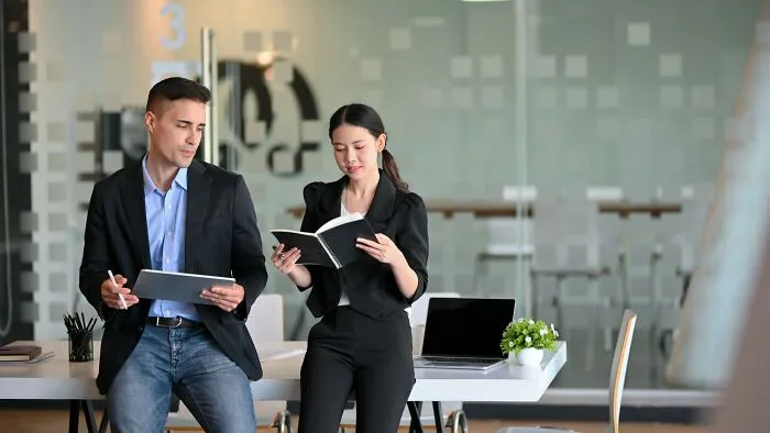 Two colleagues discussing work in a modern office, showing weird at work behaviors with a tablet and notebook