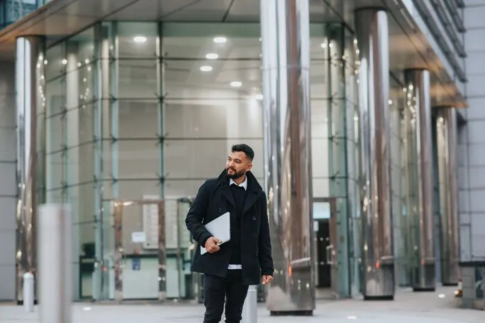Professional man holding a laptop outside a modern glass office building, looking weird at work in formal wear.