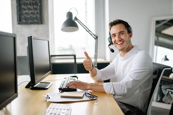 Smiling office employee with a headset giving a thumbs up, looking weird at work at his computer desk.