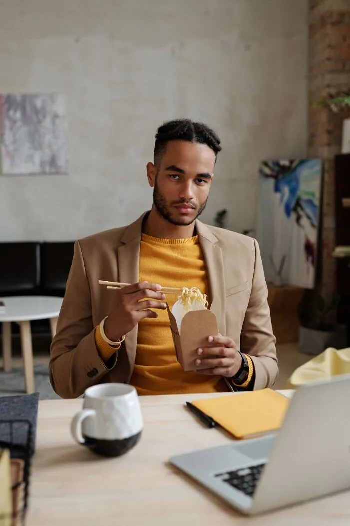 A man eating noodles at his desk, behaving weird at work while sitting in front of a laptop.