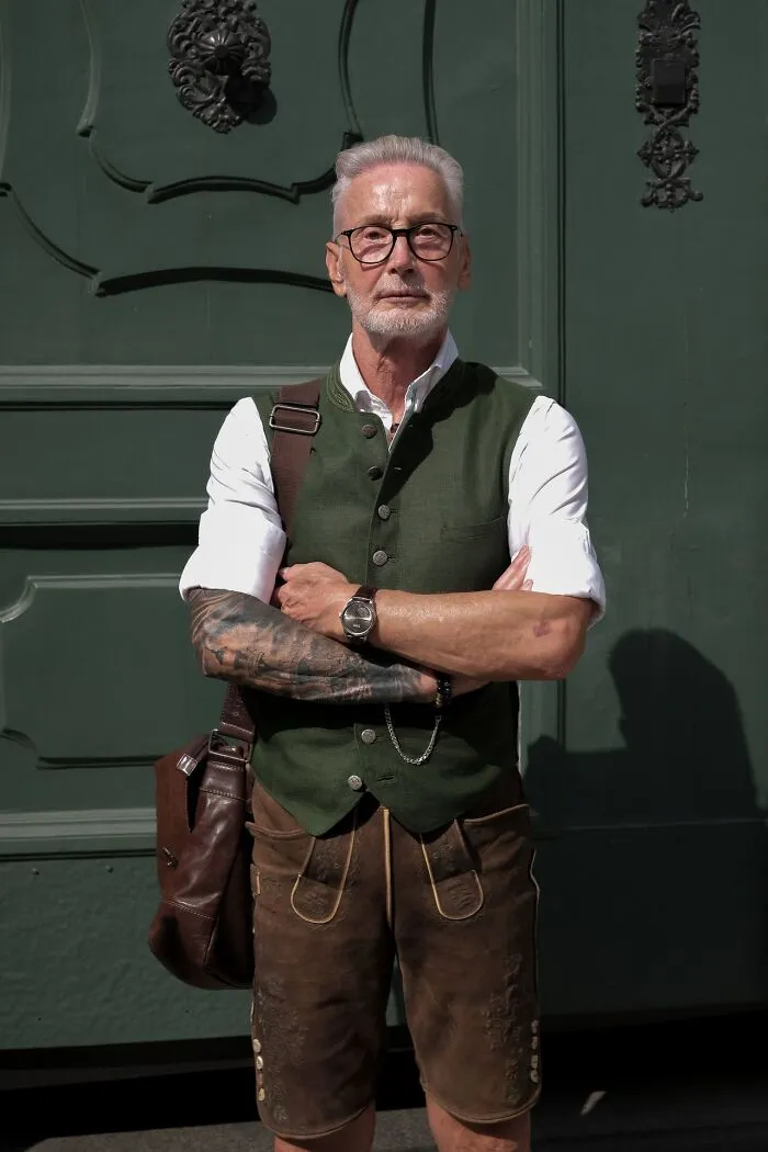 Elderly man wearing traditional Bavarian lederhosen posing outside, looking weird at work in unique attire.